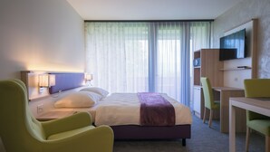 Ein Hotelzimmer mit einem Bett, zwei Stühlen, einem Schreibtisch, einem Fernseher und einem fenster mit Vorhängen.
