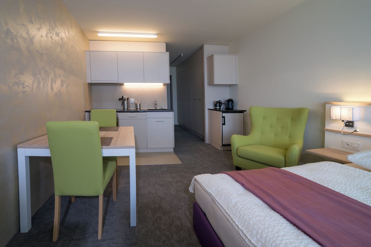 Une chambre d’hôtel compacte comprenant un coin cuisine, un espace repas avec une table et des chaises, un lit avec une tête de lit violette et une table de chevet avec une lampe.