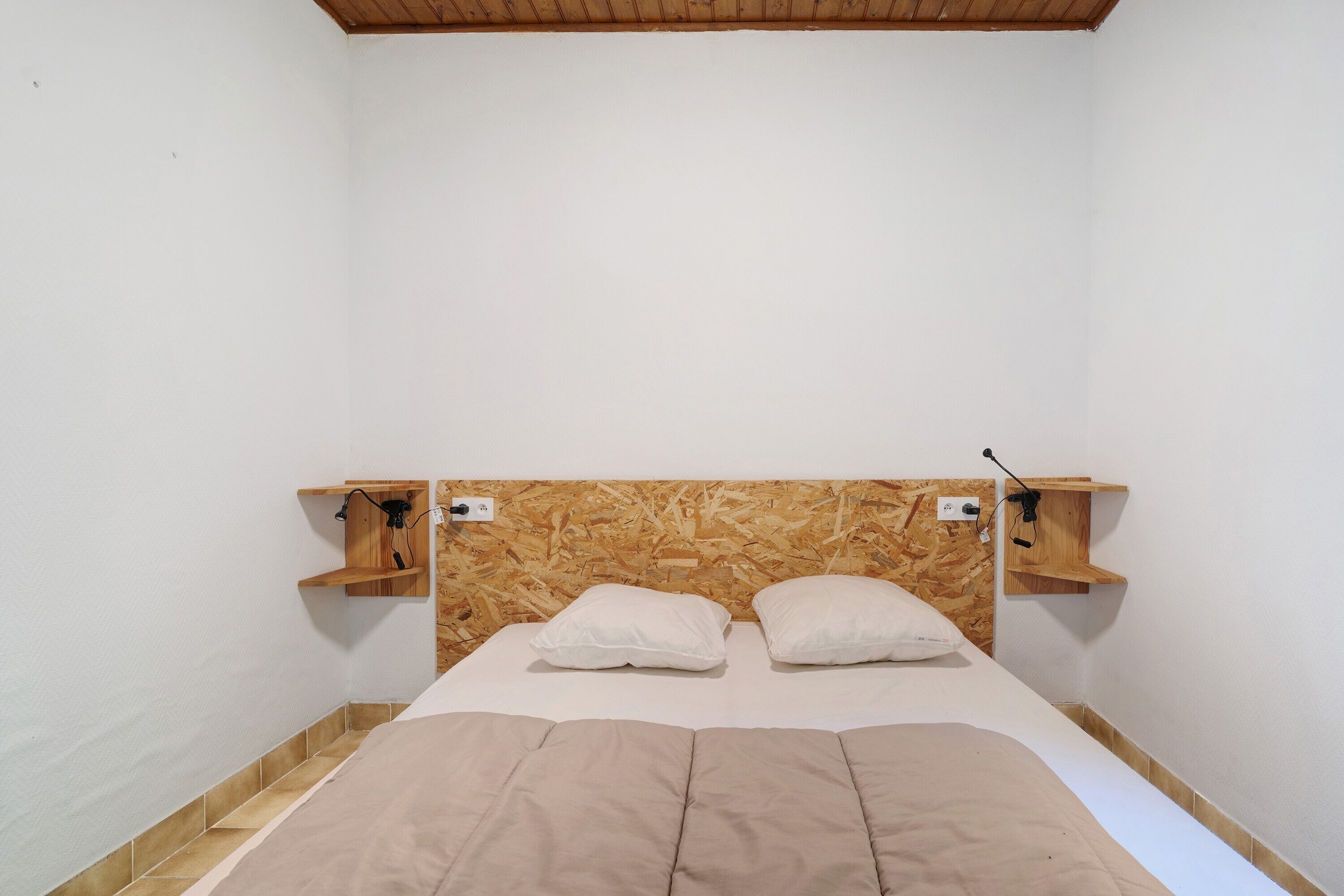 Un dormitorio con cabecera de madera, dos mesitas de noche, una cama con colcha color beige y suelo de baldosas.