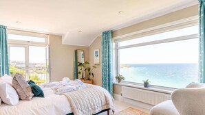 A bedroom with a bed, a chair, a large window with a view of the sea, and a radiator. - 3 Bed in St. Ives (oc-sims01) (Carbis Bay)