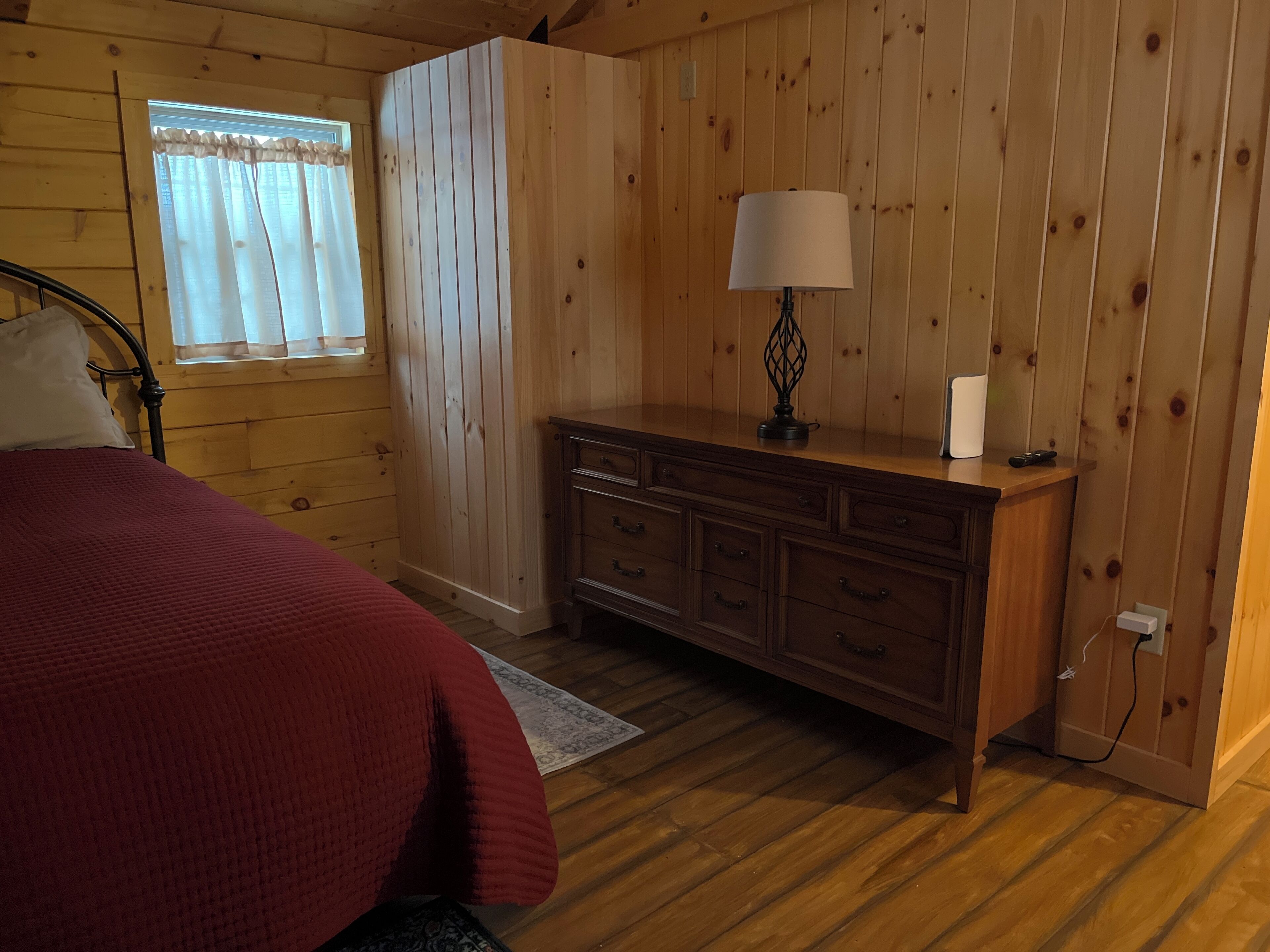 Une chambre à coucher aux murs lambrissés, avec un lit, une commode, une lampe et une fenêtre avec des rideaux.