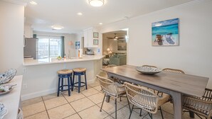 Una cucina moderna con zona pranzo, con un tavolo di legno e sedie in vimini, un'ampia finestra e un quadro a tema spiaggia.