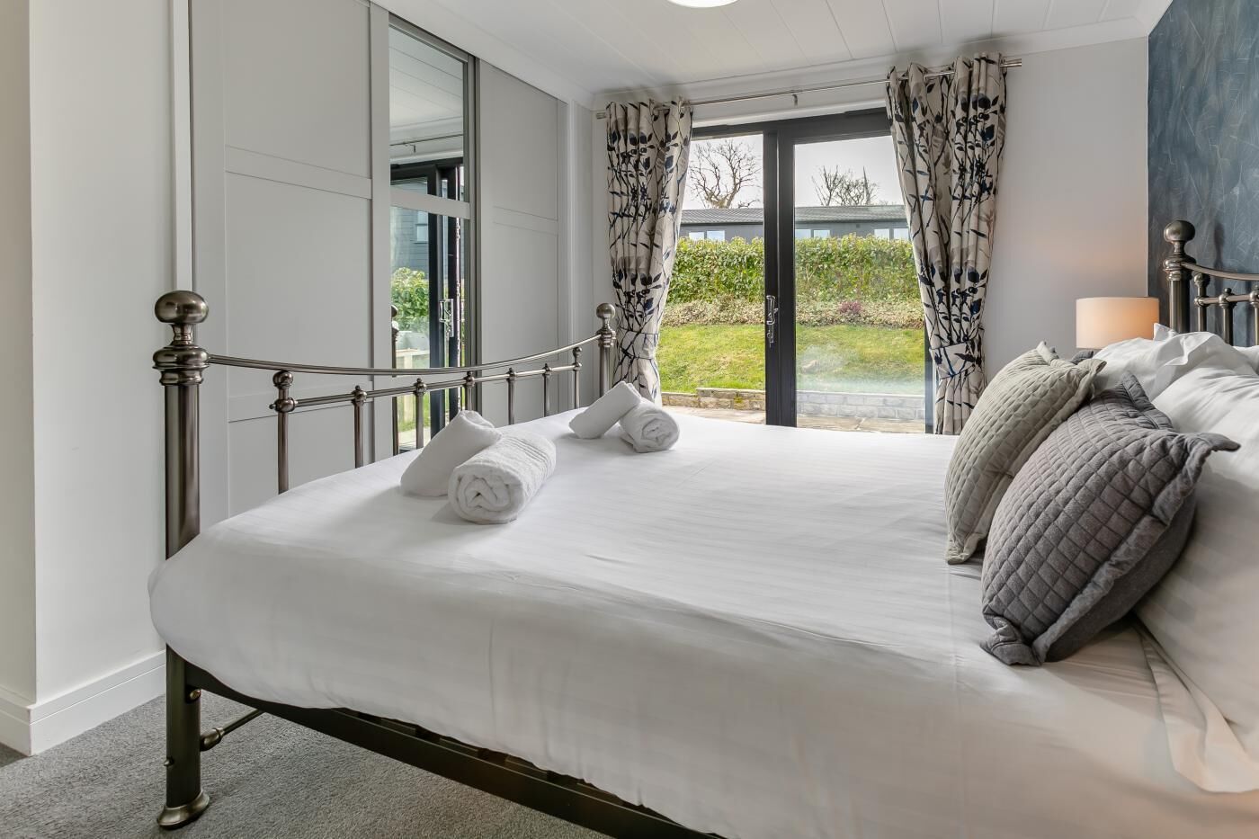 A neatly made bed with rolled towels and pillows, a metal-framed bed, and a view of greenery through a large window.