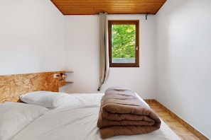 Ein Schlafzimmer mit hölzernem Kopfteil, einem Fenster mit Blick auf Grünflächen und einem Bett mit weißer und brauner Bettwäsche. Der Boden ist aus Holz.