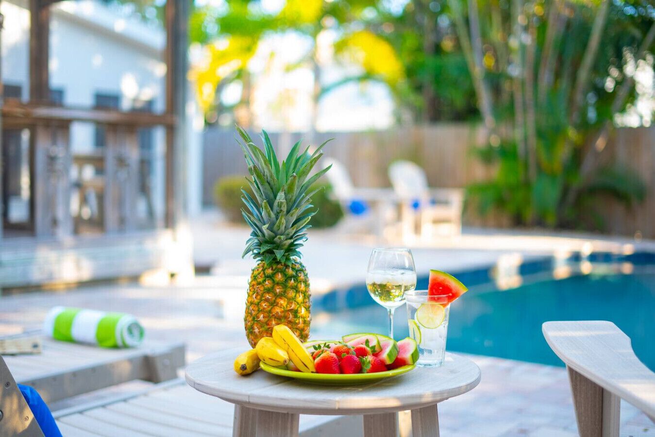 A poolside table with a pineapple, bananas, strawberries, watermelon, and a glass of white wine.