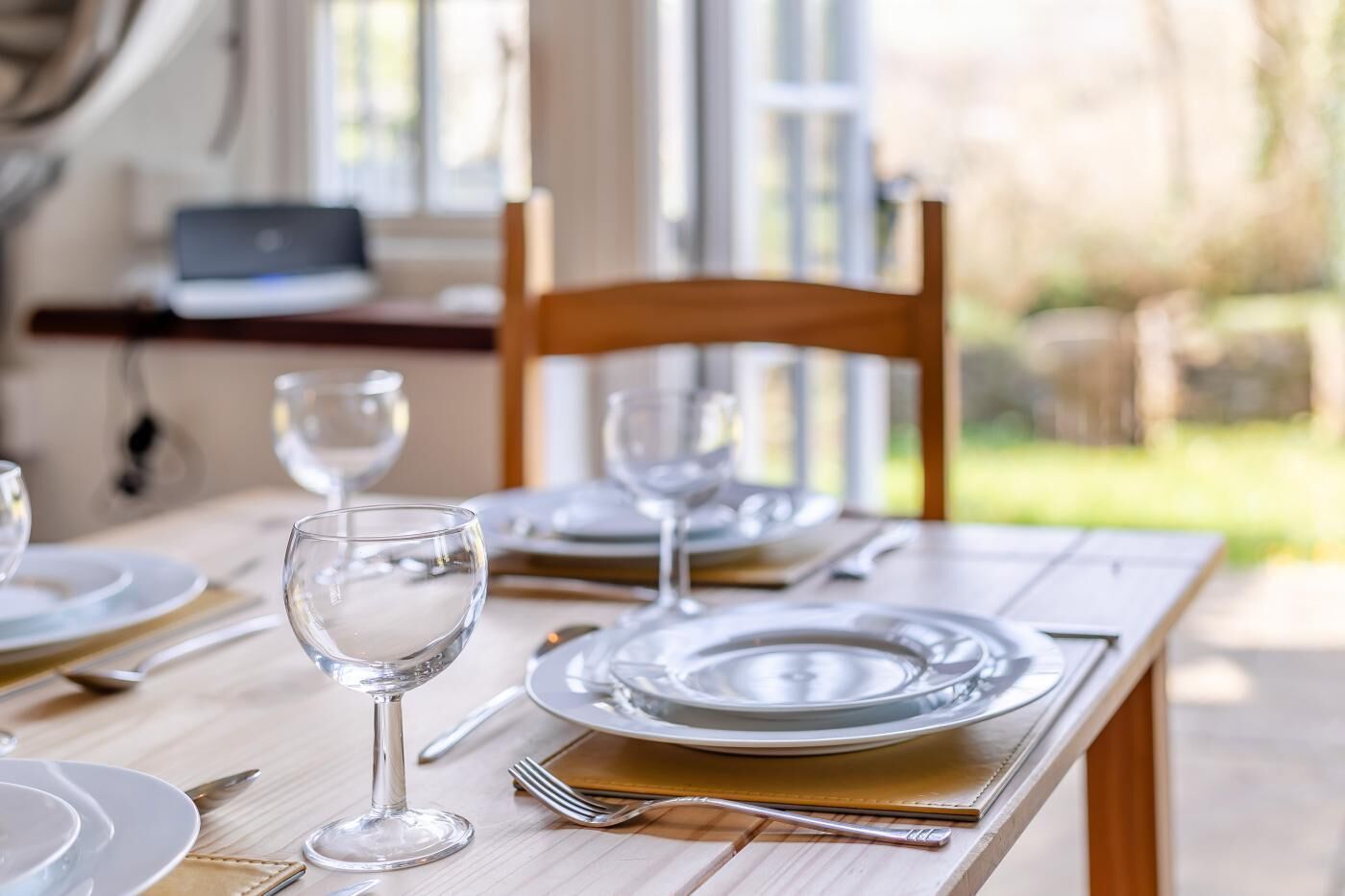 A dining table set with plates, glasses, and cutlery, with a laptop on a desk in the background.