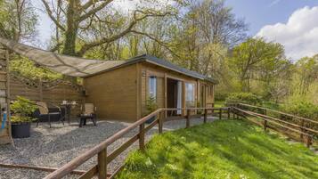 A wooden cabin with a gravel patio, a wooden deck, and a view of the surrounding forest.