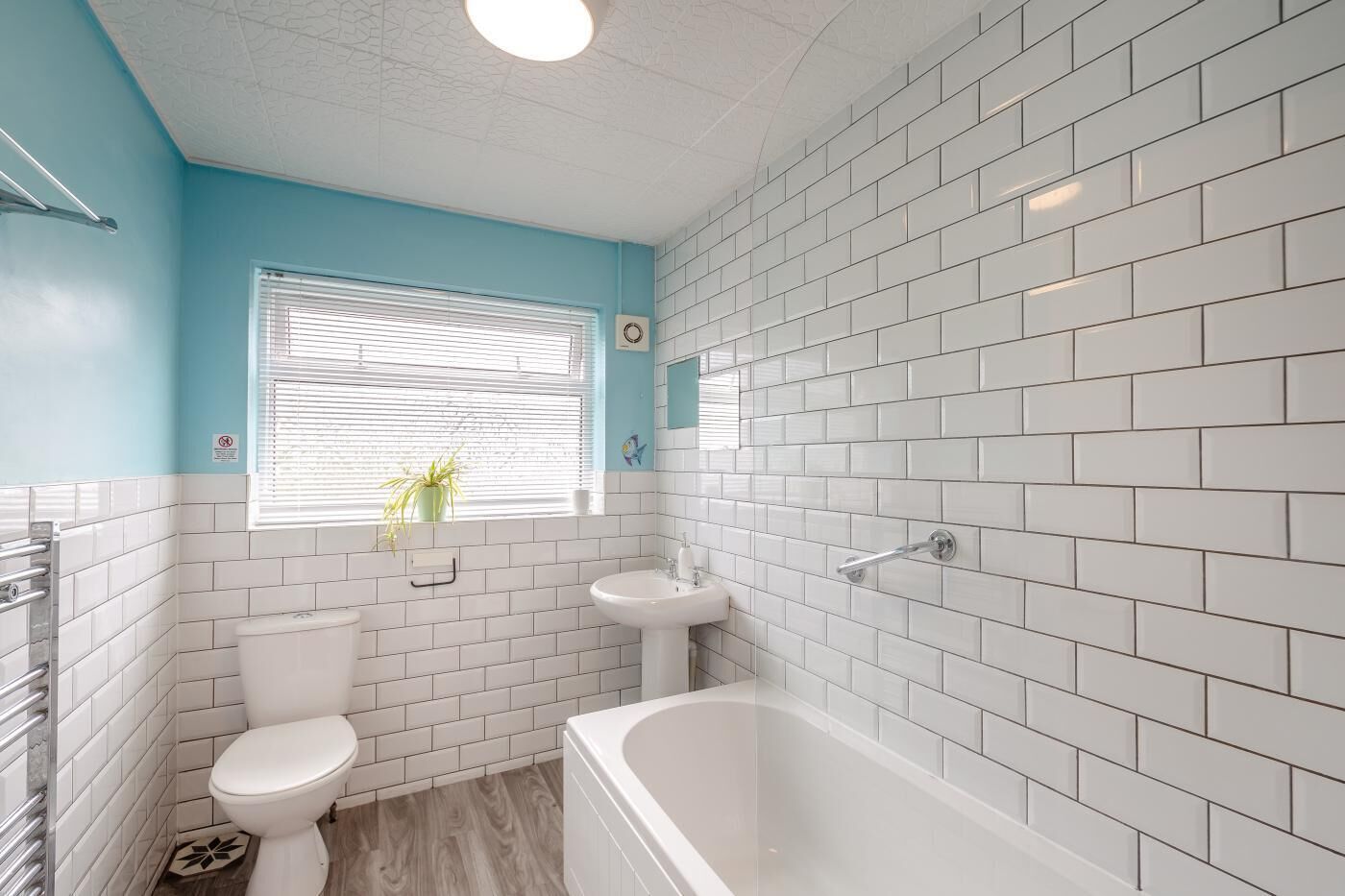 A bathroom with white subway tiles, a white bathtub, a white toilet, and a window with blinds.