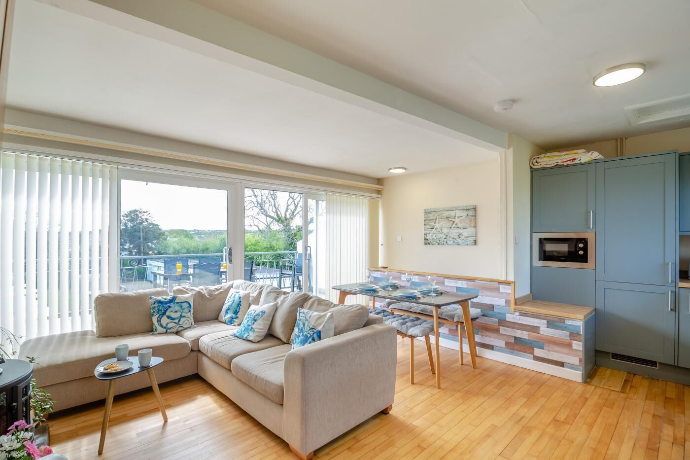 Uma sala moderna com um sofá bege, uma mesa pequena e uma área de jantar com um banco de madeira. Há também uma cozinha com micro-ondas e armários.