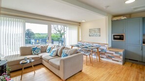 Ein modernes Wohnzimmer mit einem beigen Sofa, einem kleinen Tisch und einem Essbereich mit einer hölzernen Bank. Es gibt eine Küchenzeile mit einer Mikrowelle und Schränken.