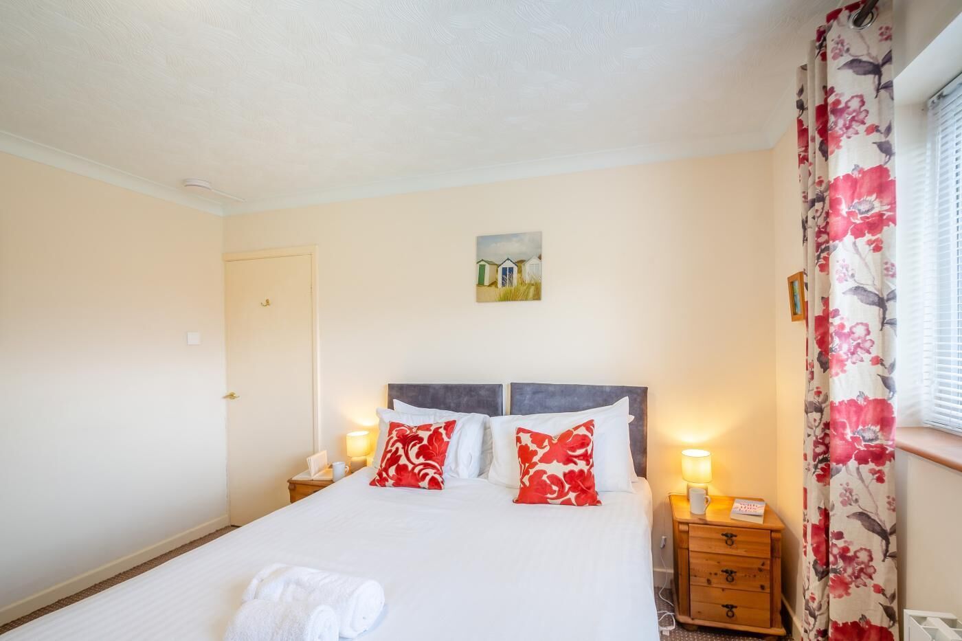 Ein ordentlich bezogenes Bett mit weißer Bettwäsche und roten und weißen Kissen, ein Nachttisch mit Lampe und ein Fenster mit blumigem Vorhang.