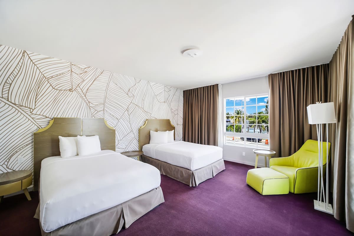A hotel room with two beds, a yellow armchair, a small table, and a window with a view of palm trees.
