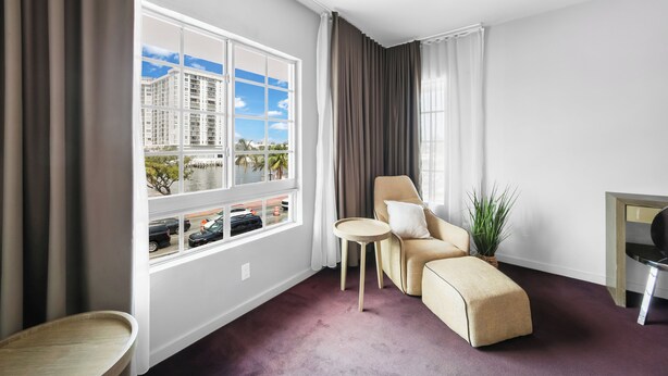 Moderni hotellihuone, jossa on suuri ikkuna, vaaleanbeige sohva, pieni pyöreä pöytä ja ruukkukasvi.