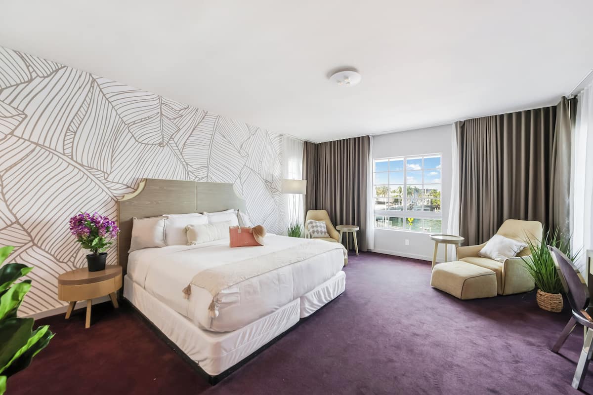 A hotel room with a large bed, a chair, a small table, and a view of the outdoors.
