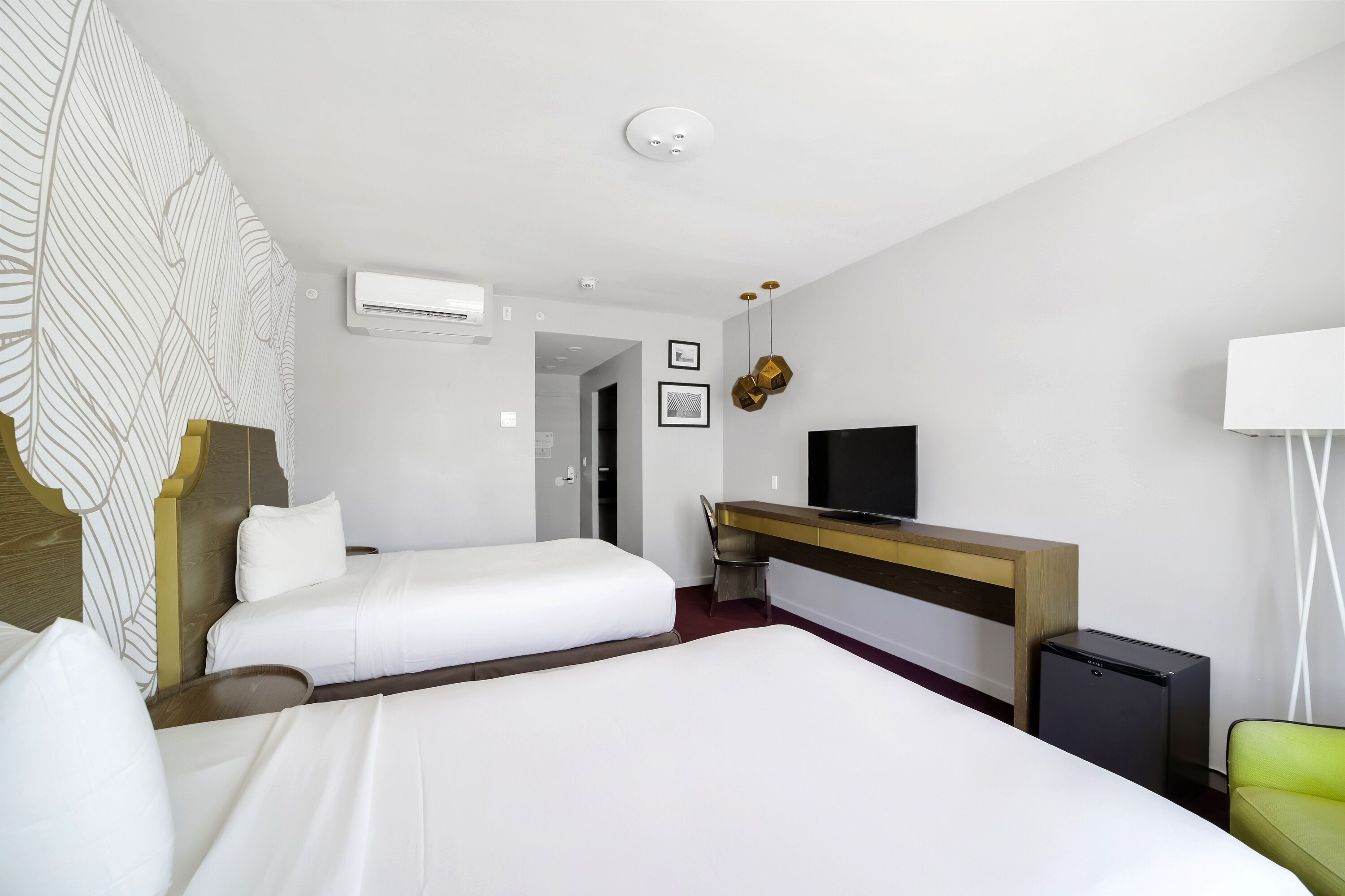 Una habitación de hotel con dos camas, un escritorio con televisor, un equipo de aire acondicionado y un refrigerador pequeño.