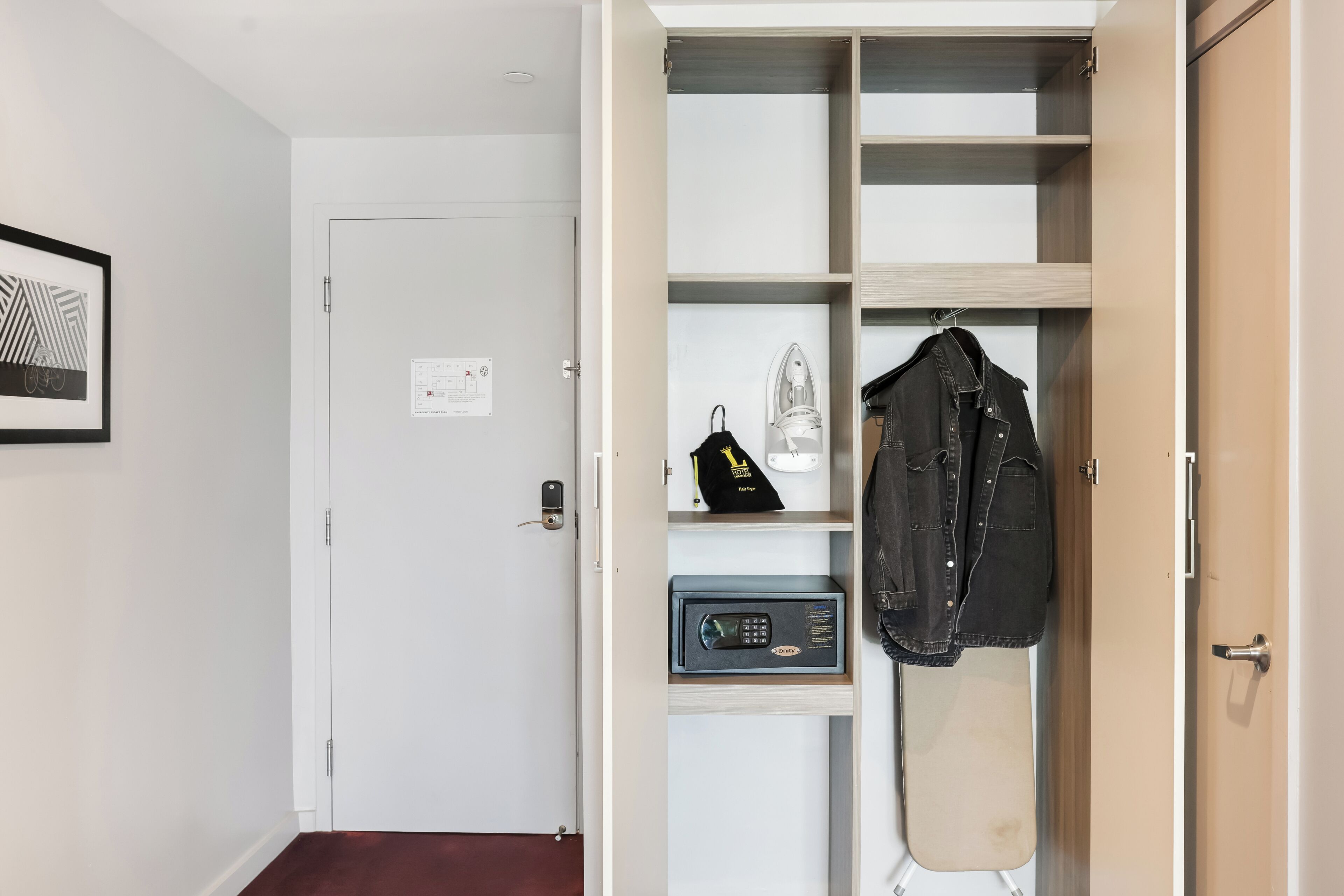 Ein Zimmer mit weißer Tür, einem gerahmten Bild an der Wand, einem Kleiderhaken mit Jacke, einem kleinen Safe und einem Regal mit einer Tasche.