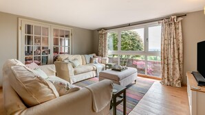 Una sala de estar con un sofá, una mesa de centro y un televisor. Hay una ventana con cortinas y un balcón con plantas.