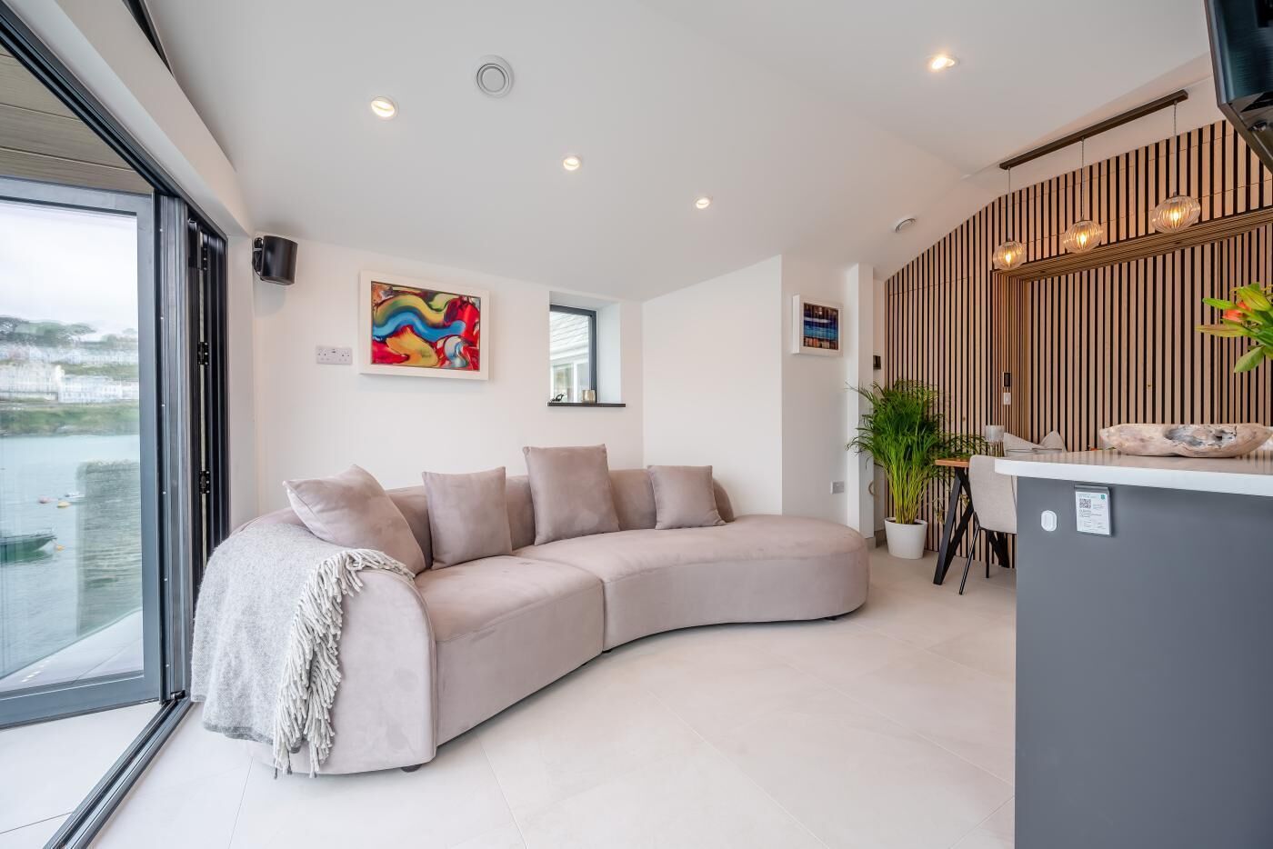 A modern living room with a curved sofa, a small bar area, and a large window overlooking a body of water.