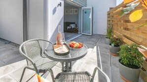 Een terras met twee rieten stoelen, een tafel met een fles en glazen, en een schaal fruit.