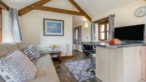 A cozy living room with a sofa, a small table with a vase, and a kitchen area with a TV, bar stool, and a window. - 1 Bed in Woodford (oc-o32064) (Woodford)