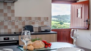 A kitchen with a view of a green landscape, a dining table set with plates and a glass of wine, and a loaf of bread on a cutting board. - 1 Bed in Woodford (oc-o32064) (Woodford)