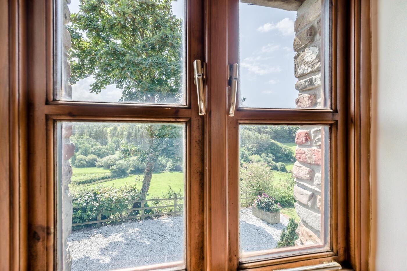 Une fenêtre à cadre en bois, dotée de deux poignées, qui offre une vue sur un jardin luxuriant avec un arbre et un mur de pierre.