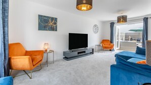 Ett modernt vardagsrum med en platt-TV, orange och blå soffor, ett litet sidobord med en lampa och ett stort fönster med gardiner.