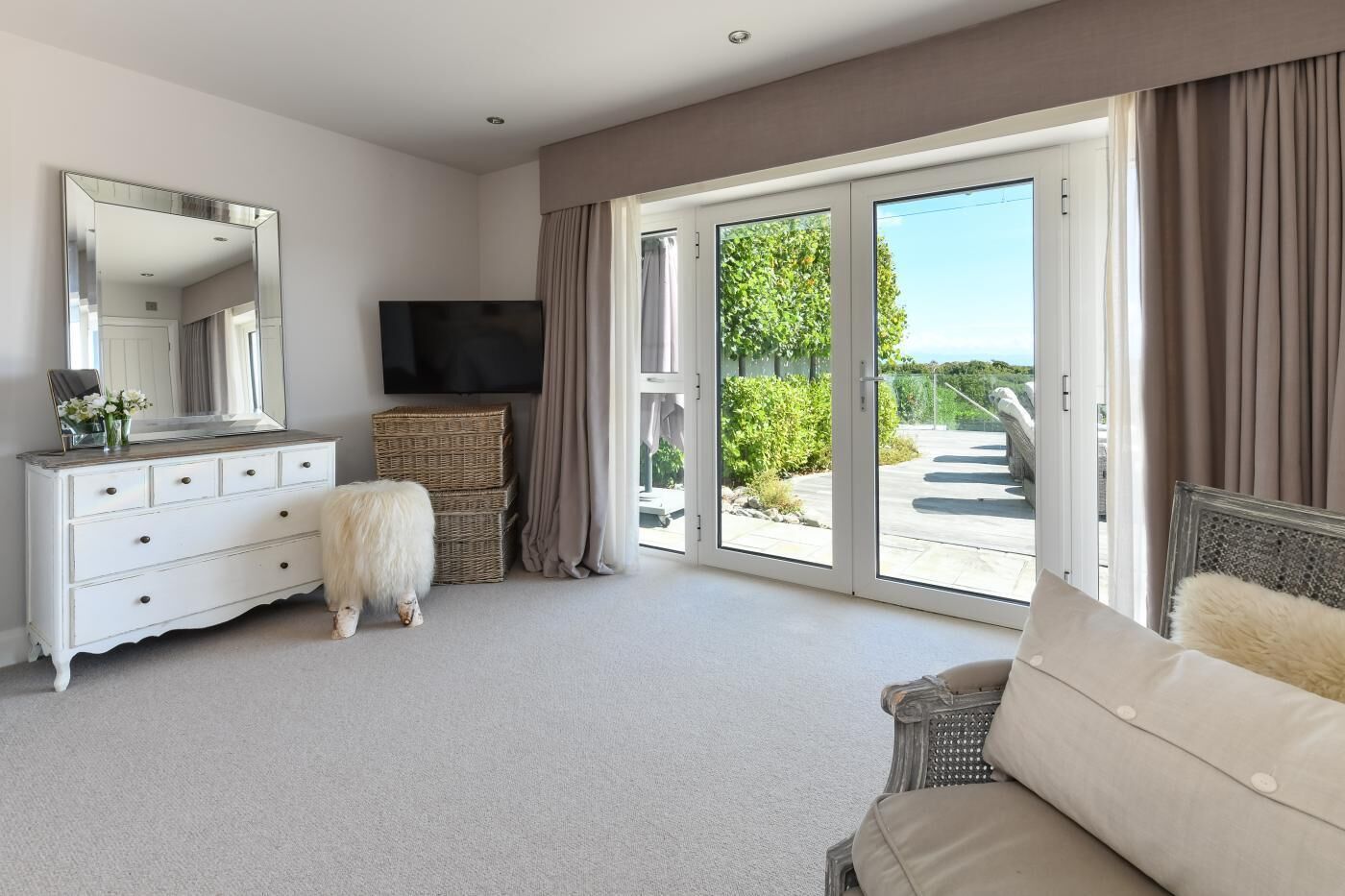 A spacious room with a large sliding glass door leading to a balcony, a white dresser with drawers, a television, and a fluffy white dog.