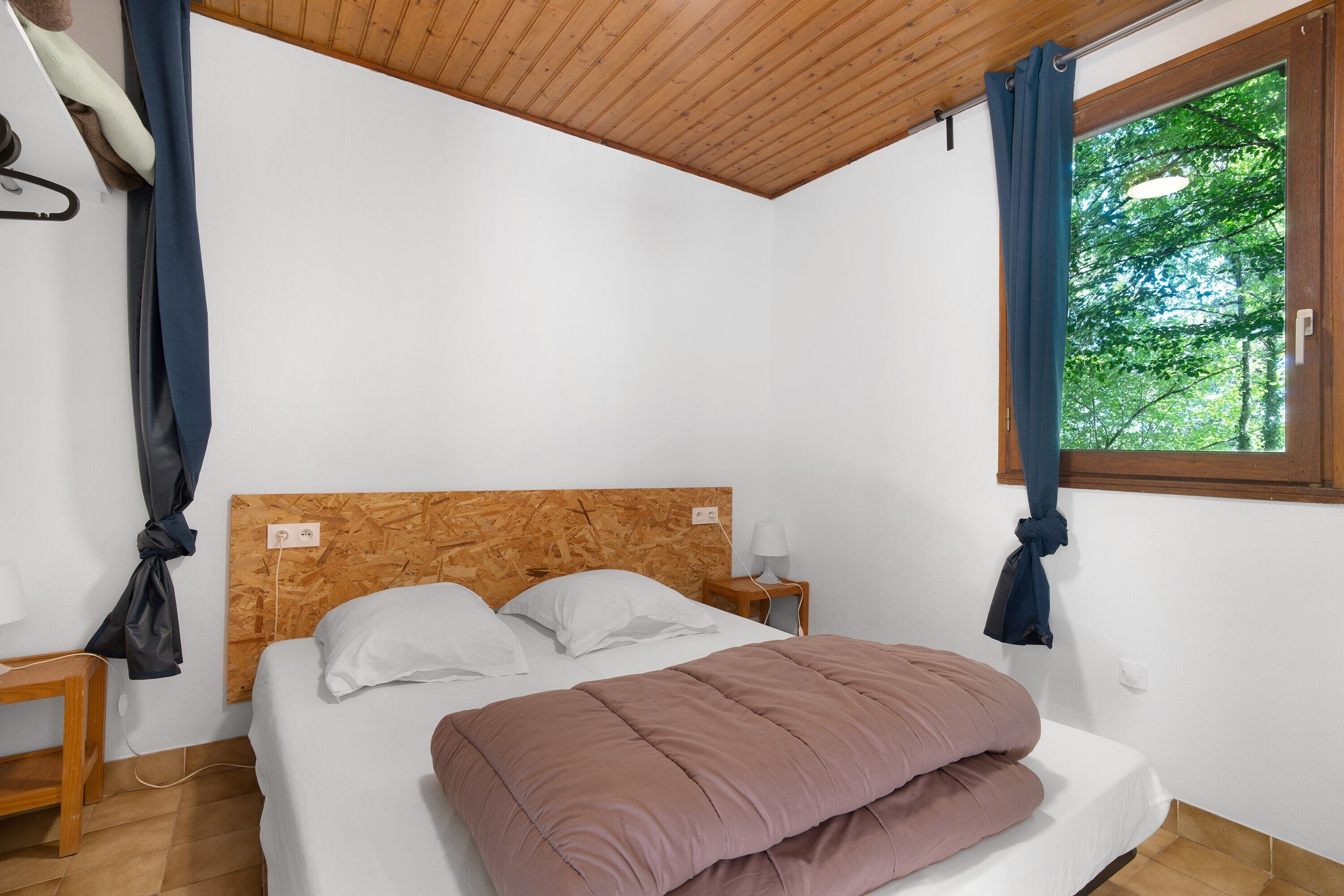 Ein Schlafzimmer mit hölzernem Kopfteil, einem Bett mit pinker Decke, einem Nachttisch mit Lampe und einem Fenster mit Vorhängen.