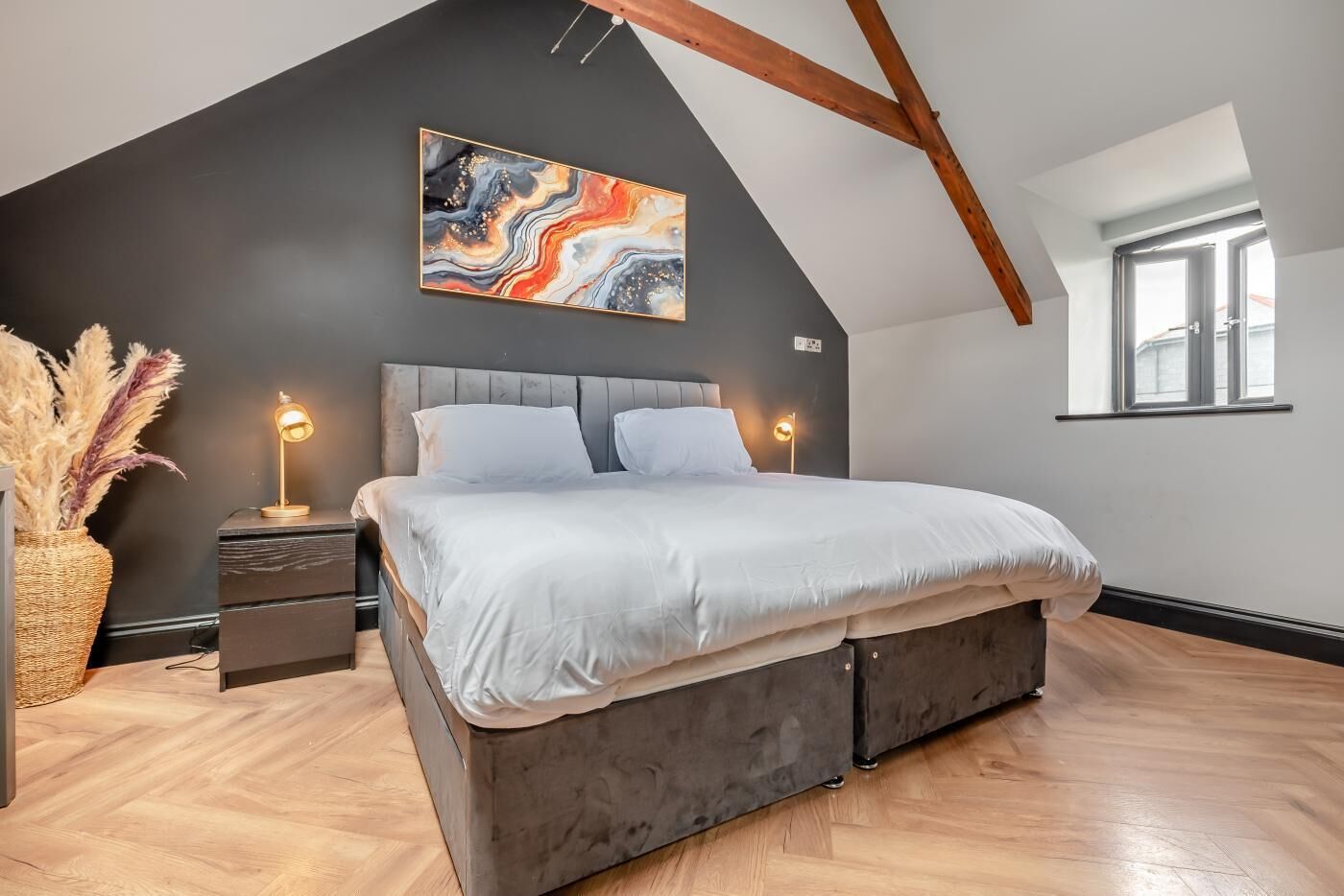 Une chambre à coucher avec un lit, des tables de chevet, une lampe, un vase contenant des plantes séchées et un tableau au mur.