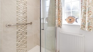 Un baño con mampara de vidrio, zona de ducha con azulejos, una ventana con cortinas de motivos florales y un adorno con forma de pez en el alféizar.