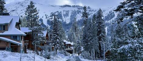 A snow-covered mountain road with buildings on either side, including a lodge with a wooden facade and a stone bridge.