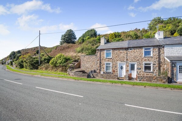A stone house with a paved road in front, surrounded by greenery and hills.