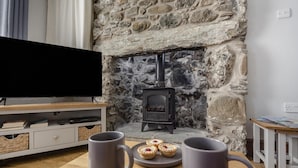 A cozy living room with a stone fireplace, a flat-screen TV, and a table with mugs and pastries. - 2 Bed in Pwllheli (oc-s32070) (Pwllheli)