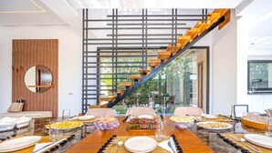 Ein zeitgemäßes Esszimmer mit einem großen Holztisch, gedeckt für sechs Personen, einer Treppe und Blick ins Freie.