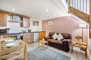 A modern kitchen with wooden cabinets, a dining area with a table and chairs, a brown leather sofa, and a small side table with a vase of flowers. - 1 Bed in Berwick-upon-Tweed (oc-a32026) (Kyloe)