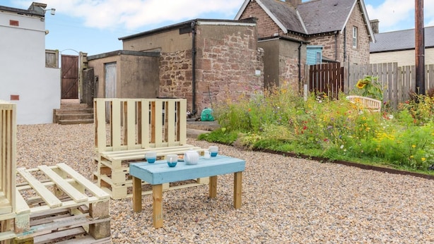 Un'area salotto esterna dallo stile rustico con tavolo e panche di legno, circondata da un vialetto sterrato e un giardino con diverse piante e fiori.