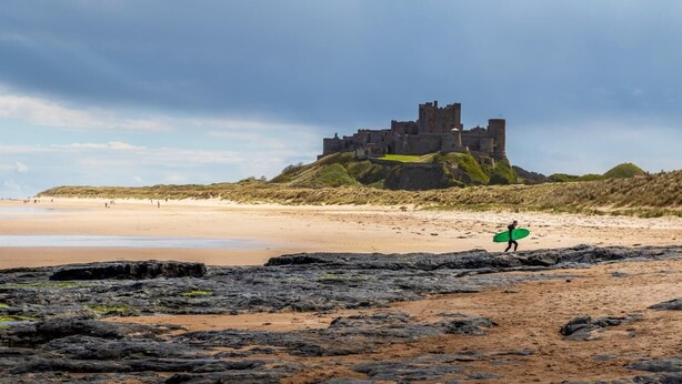A castle on a hill overlooking a beach with a person carrying a surfboard.