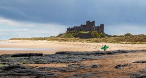 A castle on a hill overlooking a beach with a person carrying a surfboard. - 1 Bed in Berwick-upon-Tweed (oc-c32028) (Kyloe)
