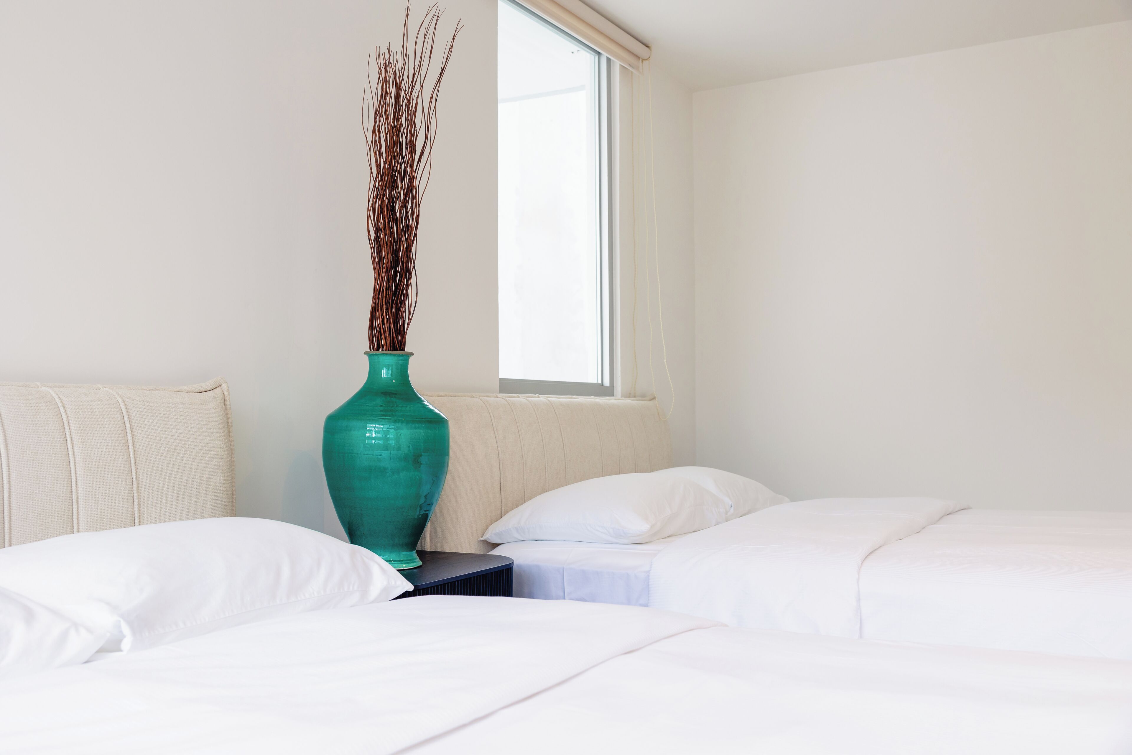 A hotel room with two beds, a teal vase with branches, and a window with blinds.