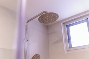 Une douche avec un pommeau de douche à main et un pommeau de douche fixe, installée dans un coin d’une pièce avec une fenêtre.