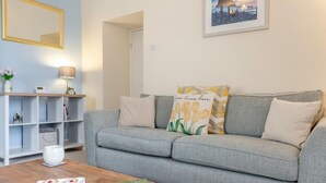 Ein Wohnzimmer mit grauem Sofa, einem Couchtisch mit Gebäck und einem Buch, einem Bücherregal, einer Lampe und einem gerahmten Bild an der Wand.