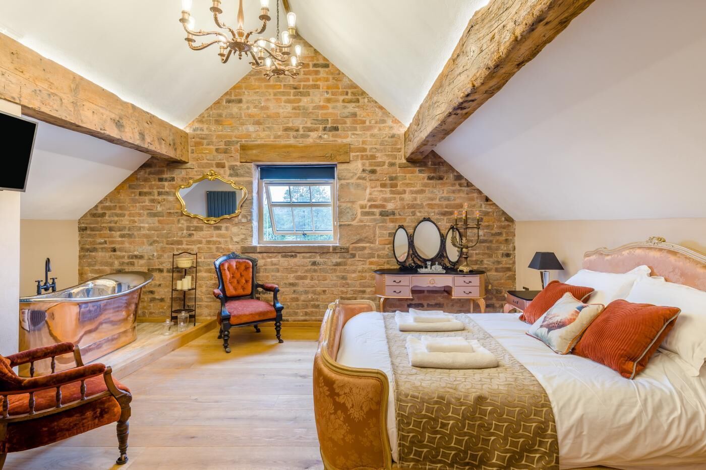 A bedroom with a sloped ceiling, a stone wall, a large bed, a vanity, a chair, and a bathtub.