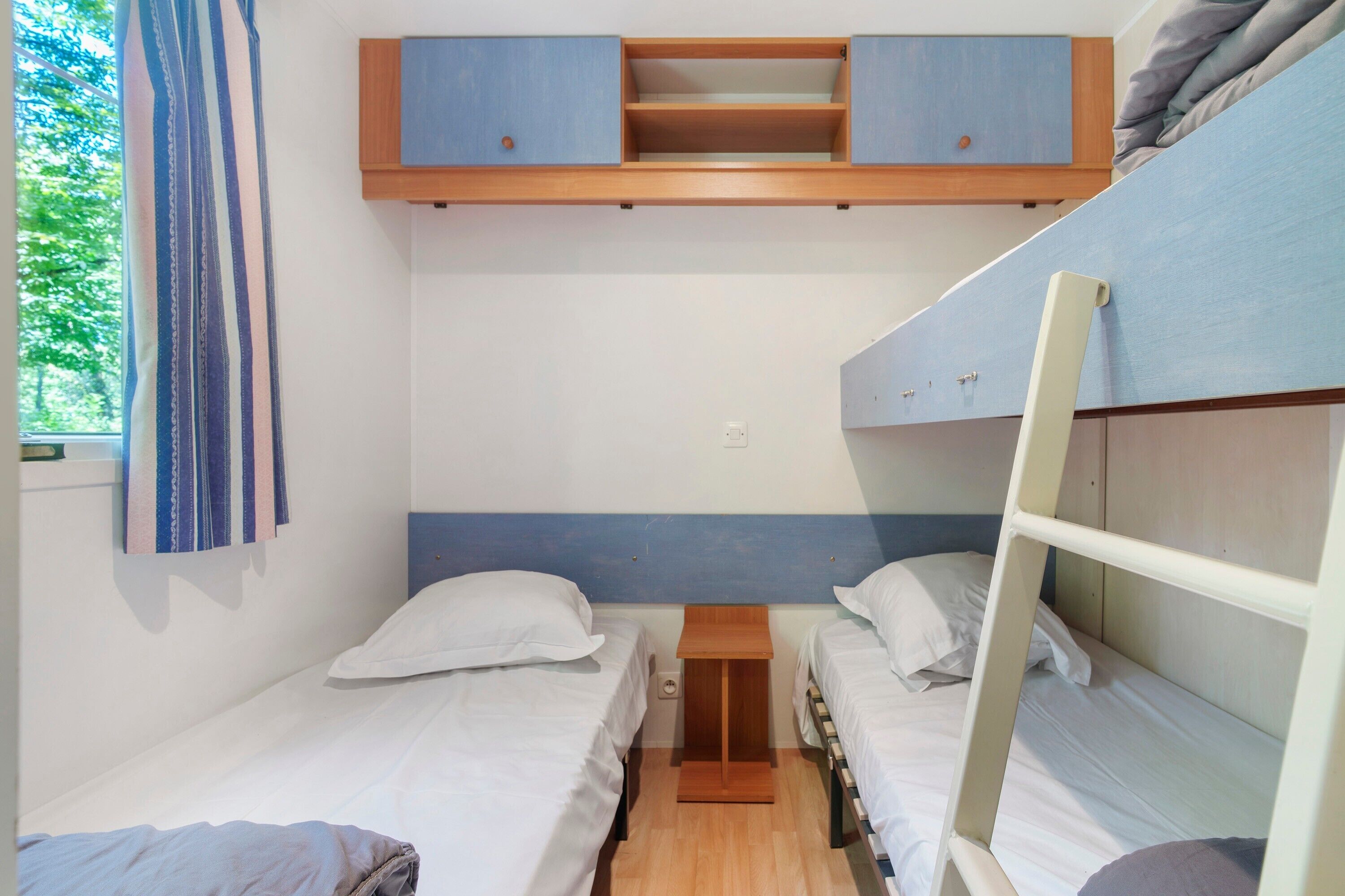 A dorm-style room with bunk beds, a small wooden nightstand, and a window with curtains.