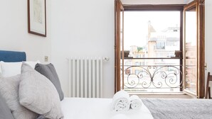 Ein Schlafzimmer mit Bett, Kissen, Heizkörper und Blick auf Gebäude durch eine offene Tür.