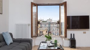 A living room with a sofa, a coffee table with decorative items, a TV, and a window with an open view of buildings. - Rental in Rome Giulio Cesare Penthouse (Rome)