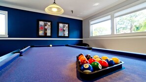 Une table de billard avec des billes, dans une pièce aux murs bleus, ornée de deux œuvres d’art encadrées, et une fenêtre donnant sur des arbres.