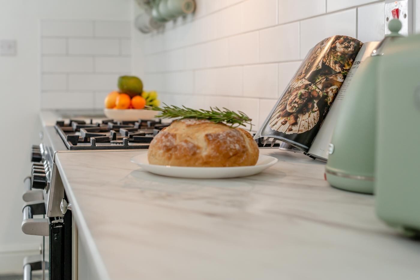 A kitchen with a loaf of bread on a plate, a fruit bowl, and a cookbook on the counter.