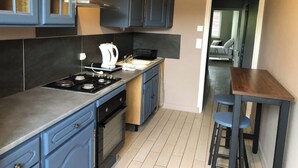 A modern kitchen with dark blue cabinets, a gas stove, an oven, and a coffee maker. There is a wooden table and two bar stools in the adjacent dining area.