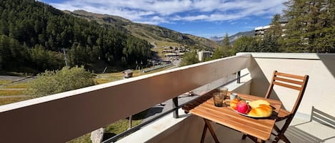 A balcony with a wooden table and chairs, overlooking a mountainous landscape with a clear sky.